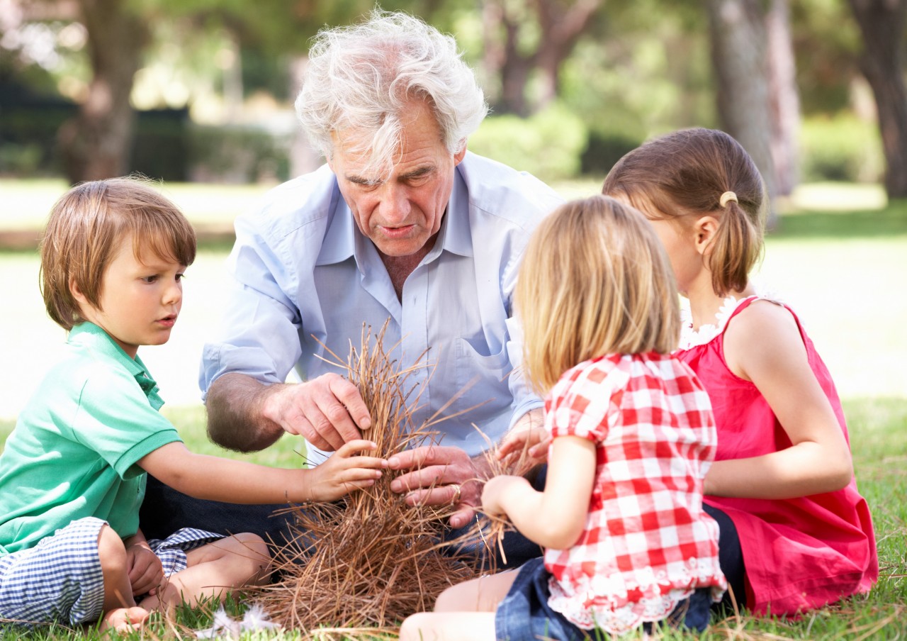 older man teaches tree young children how to build a fire with stick pile